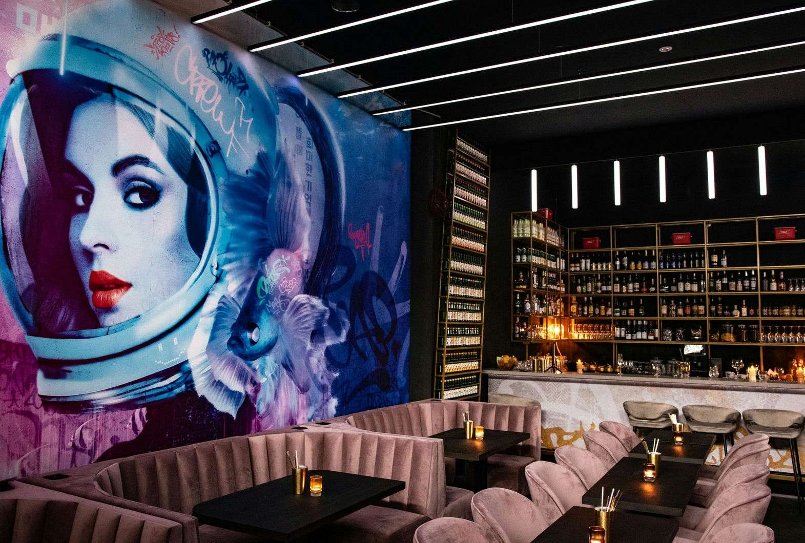 Barzaal met een muurvullend portret van een vrouw in een ruimtehelm, roze fluwelen banken ervoor en een verlichte flessenwand rechts.