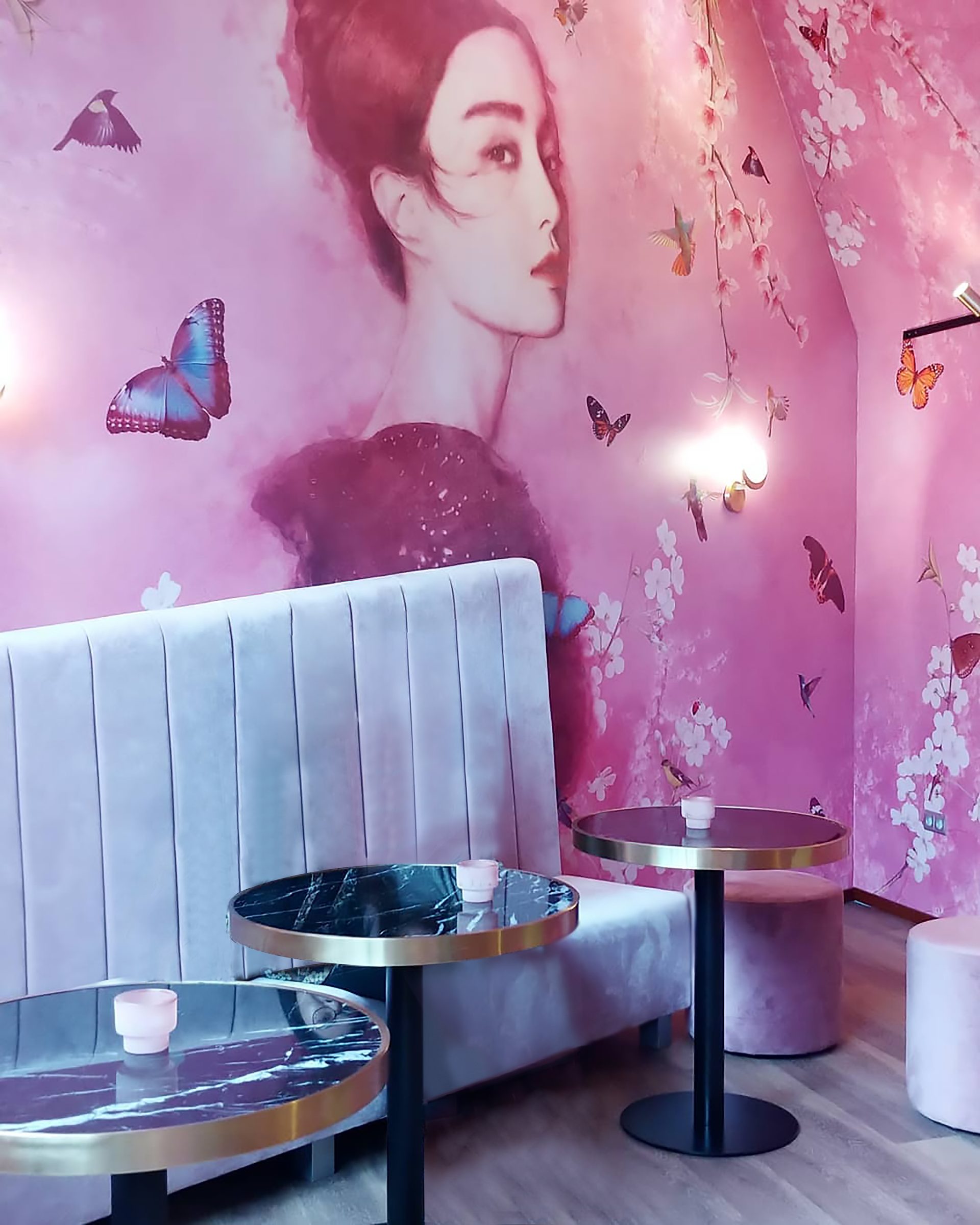 Roze interieurwand met een geschilderde vrouwenfiguur tussen bloesemtakken, vlinders en een witte pauw, boven een fluwelen bank.