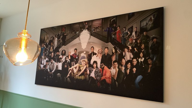 Eén breed doek aan een lichte wand, schuin gezien: een groepsportret van tientallen figuren rond een trappenhuis, met een glazen hanglamp ervoor.