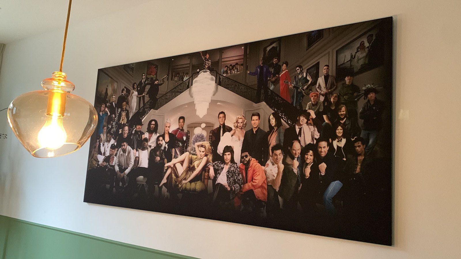 Eén breed doek aan een lichte wand, schuin gezien: een groepsportret van tientallen figuren rond een trappenhuis, met een glazen hanglamp ervoor.