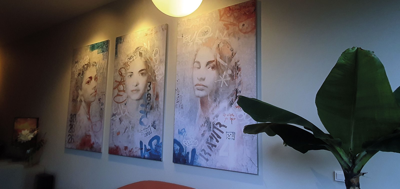 Drieluik op doek aan een woonkamerwand: drie vrouwenportretten in oplopend formaat, met witte en blauwe graffitilijnen over de gezichten, onder een bollamp.