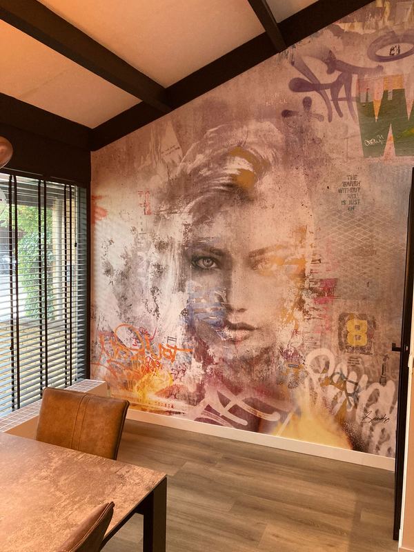 Eetkamerwand van vloer tot plafond: een vrouwengezicht in gedempt roze en grijs met oranje en paarse tags eroverheen, naast een houten tafel met leren stoel.