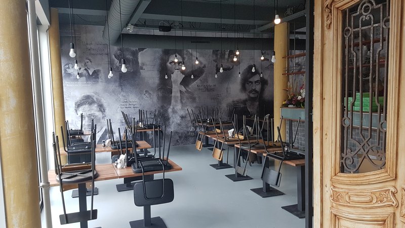 Restaurantzaal vóór opening, met stoelen op de tafels; langs de volle achterwand een grijze collage van portretten met handgeschreven tekst ertussen.