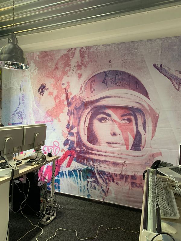 Kantoorruimte: een astronaut met een vrouwengezicht achter het vizier in roze en blauw op de achterwand, met werkplekken en monitoren ervoor.