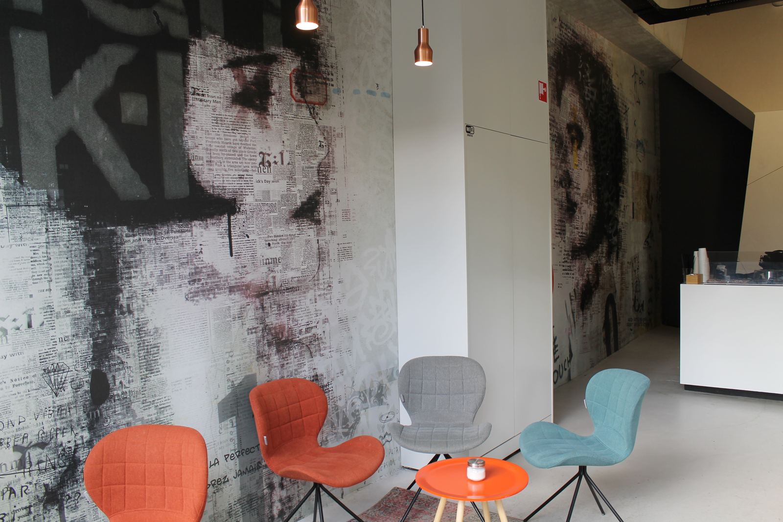 Zithoek in een horecazaak: een wandvullend werk in grijs en zwart, met een portret tussen krantenletters en graffiti, dat om de hoek van de ruimte doorloopt. Ervoor staan oranje, grijze en blauwe stoelen en een laag tafeltje.