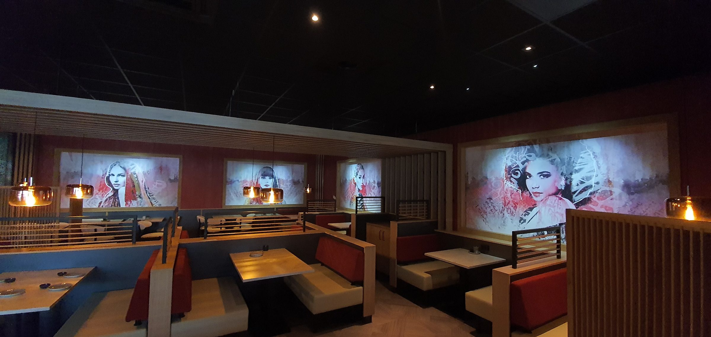 Donkere restaurantzaal waarin vier portretten in rood en grijs als verlichte panelen tussen verticale houten latten zijn geplaatst, boven booths met roodleren banken.