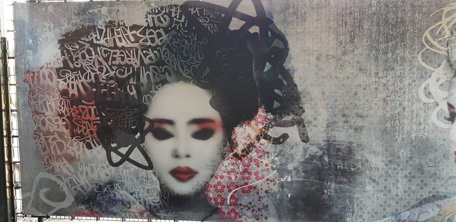 Breed muurwerk: een geishagezicht met witte make-up en rode lippen, het kapsel volledig opgebouwd uit zwarte lettertekens, op een grijze betonnen wand.