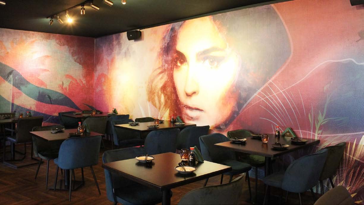 Restaurantzaal waarin een geschilderd vrouwenportret in rood, roze en oker over twee wanden doorloopt, tussen tropische bladeren. Ervoor staan gedekte tafels met donkerblauwe stoelen onder spotverlichting.