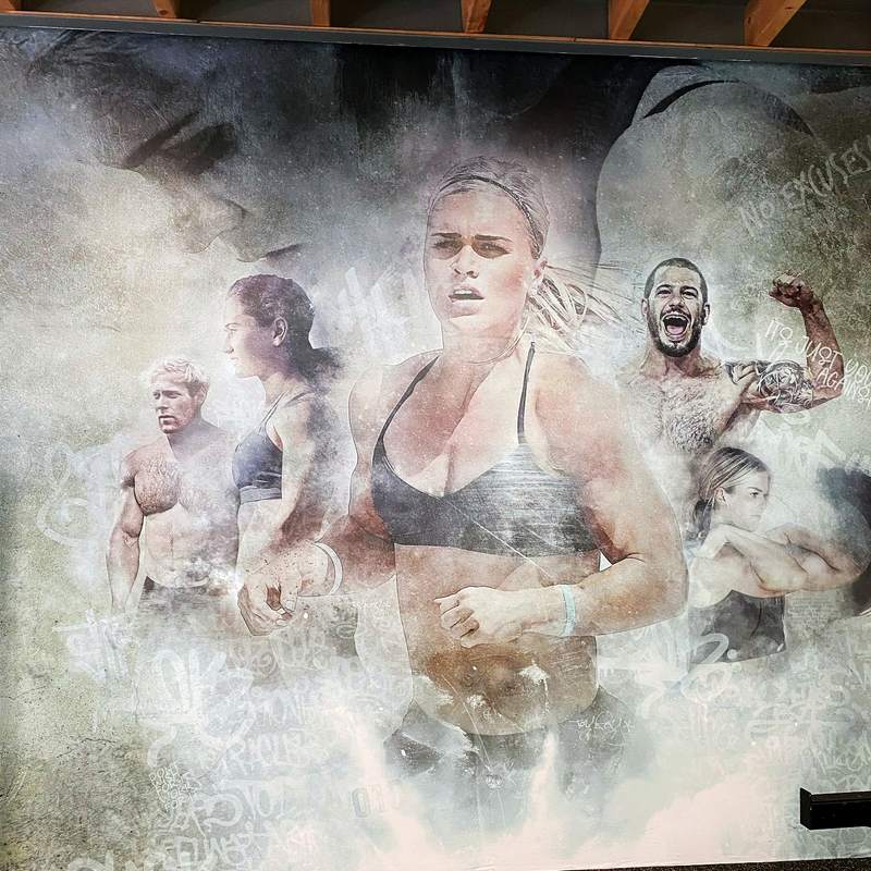 Muurvullend werk in een sportruimte: vijf sporters in grijs en okerwit, uit een wolk van stof en graffitiletters naar voren komend.