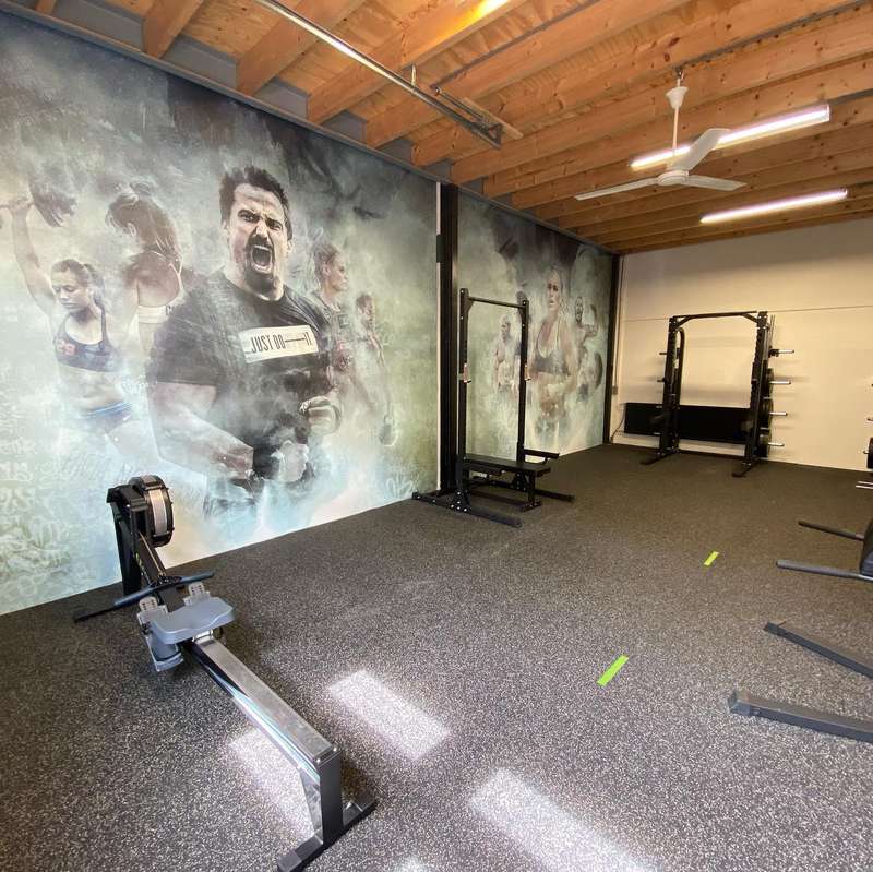Trainingszaal met open houten kap en rubberen vloer; het beeld van sporters loopt over twee wanden door, met een roeimachine en squat racks ervoor.