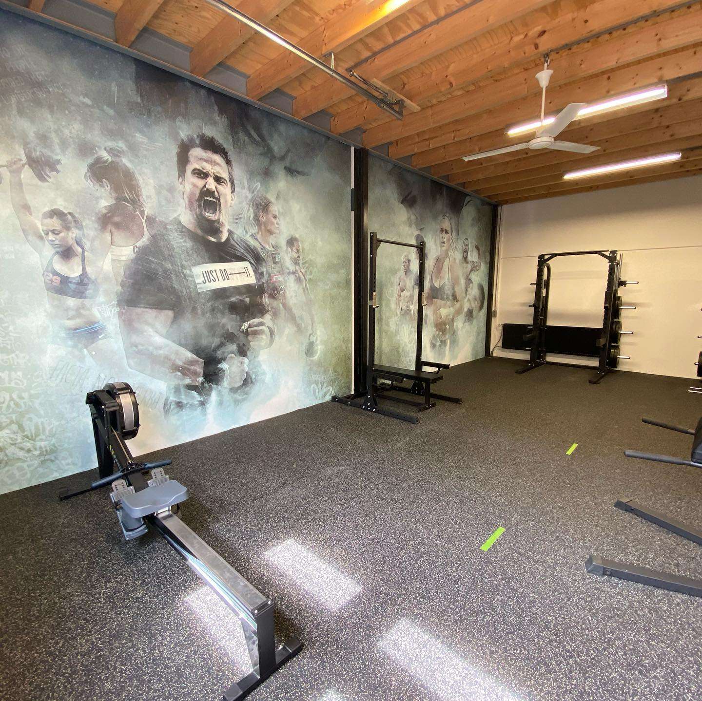 Trainingszaal met open houten kap en rubberen vloer; het beeld van sporters loopt over twee wanden door, met een roeimachine en squat racks ervoor.
