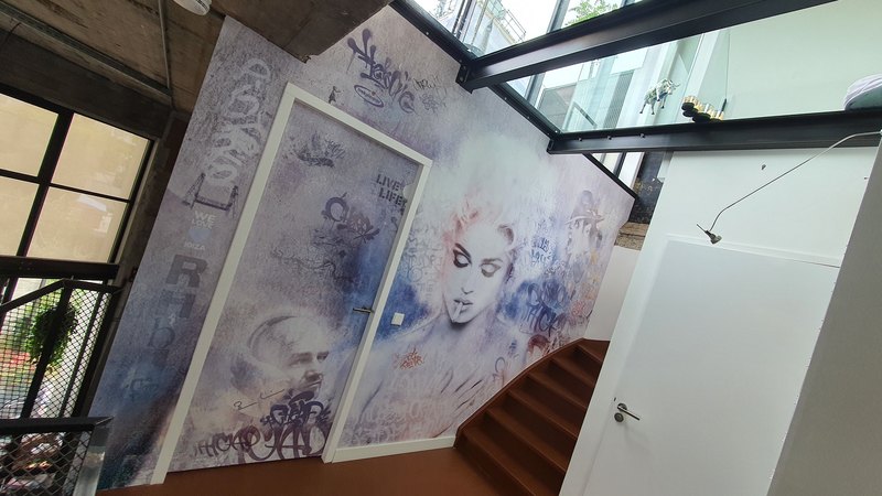 Trappenhuis onder een glazen kap met stalen liggers; de hele wand, inclusief het witte deurblad, draagt één blauwviolet beeld met een figuur en tags.