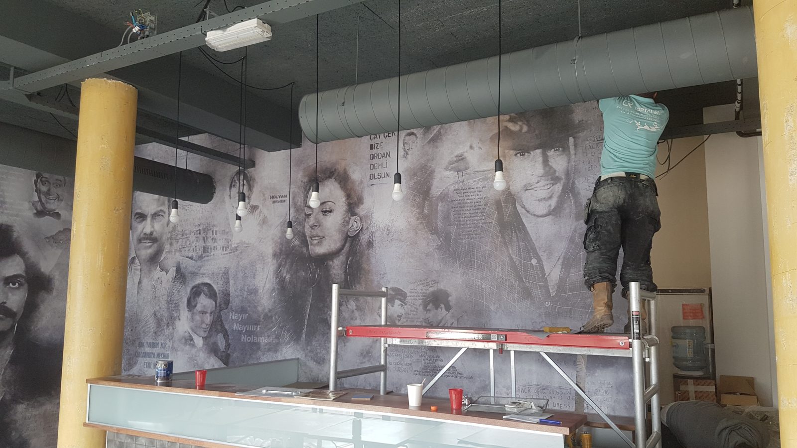 Wandvullende muurschildering met zwart-witte portretten en handgeschreven tekst in een horecaruimte; rechts staat iemand op een rolsteiger aan de wand te werken.