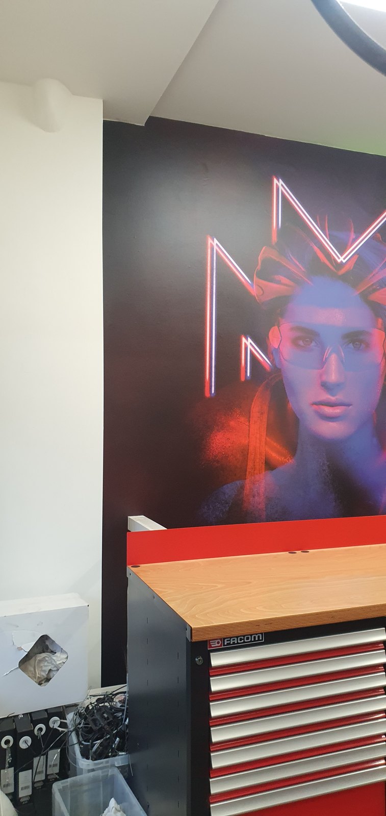 Werkruimte met een wandvullend werk over de volle hoogte: een portret in neonblauw en rood op een donkere ondergrond, achter een rode werkbank.