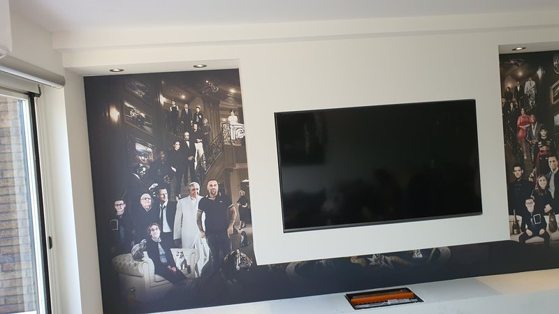 Muurvullende print rond een tv-nis: hetzelfde groepsportret in donkere tinten loopt links en rechts van het scherm door, met daglicht uit een schuifpui erlangs.
