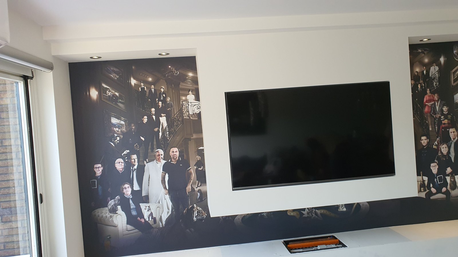 Muurvullende print rond een tv-nis: hetzelfde groepsportret in donkere tinten loopt links en rechts van het scherm door, met daglicht uit een schuifpui erlangs.